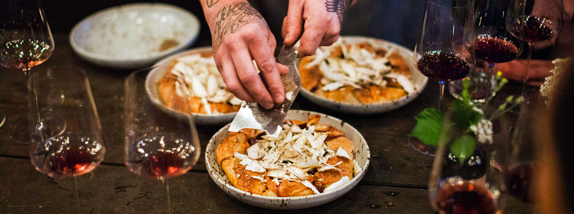 A chef puts together three dishes on a table surrounded by glasses of red wine.
