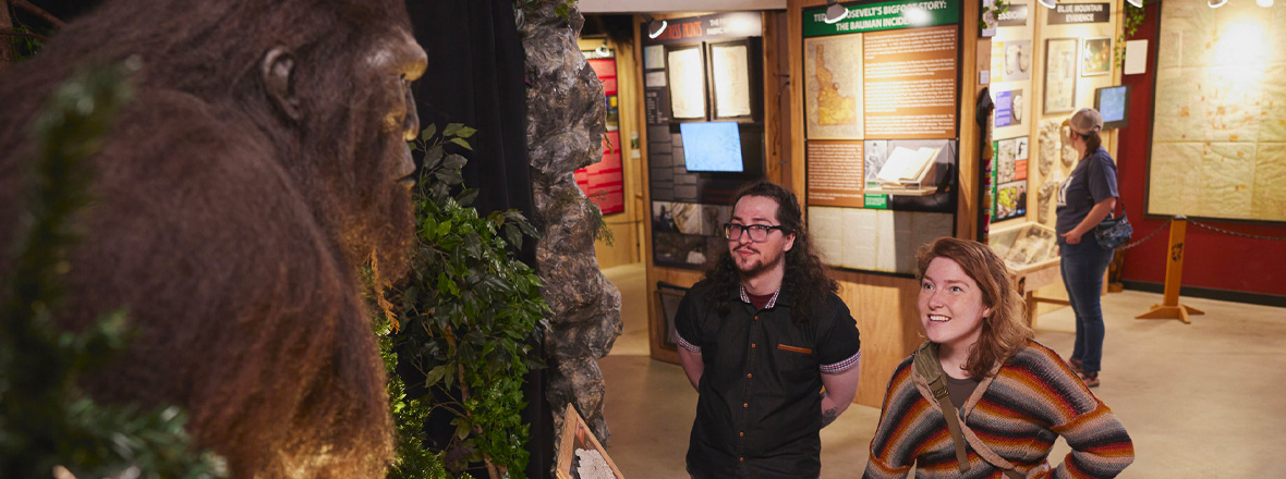 A young man and woman looking at a tall Bigfoot statue inside a museum.