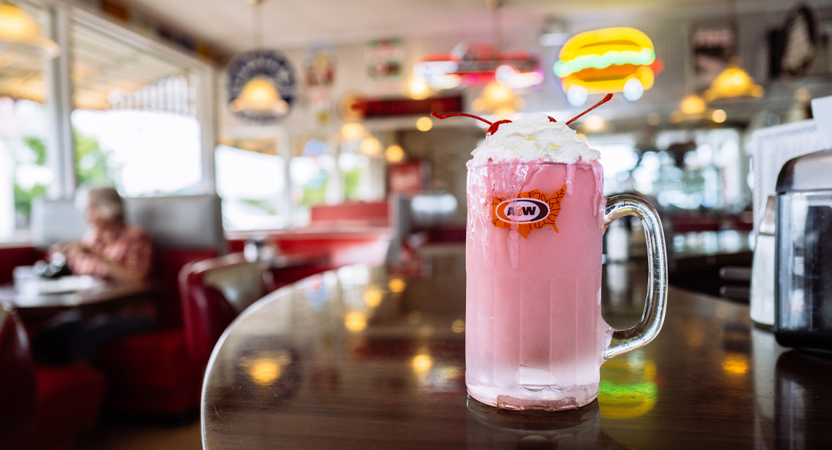 A tall glass of a frosty pink drink topped with whipped cream on a diner counter.
