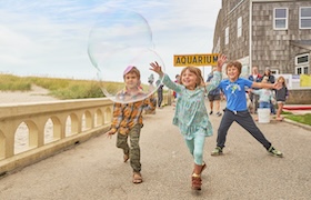 Children run along a seaside walkway blowing a large bubble, with an aquarium building in the background.