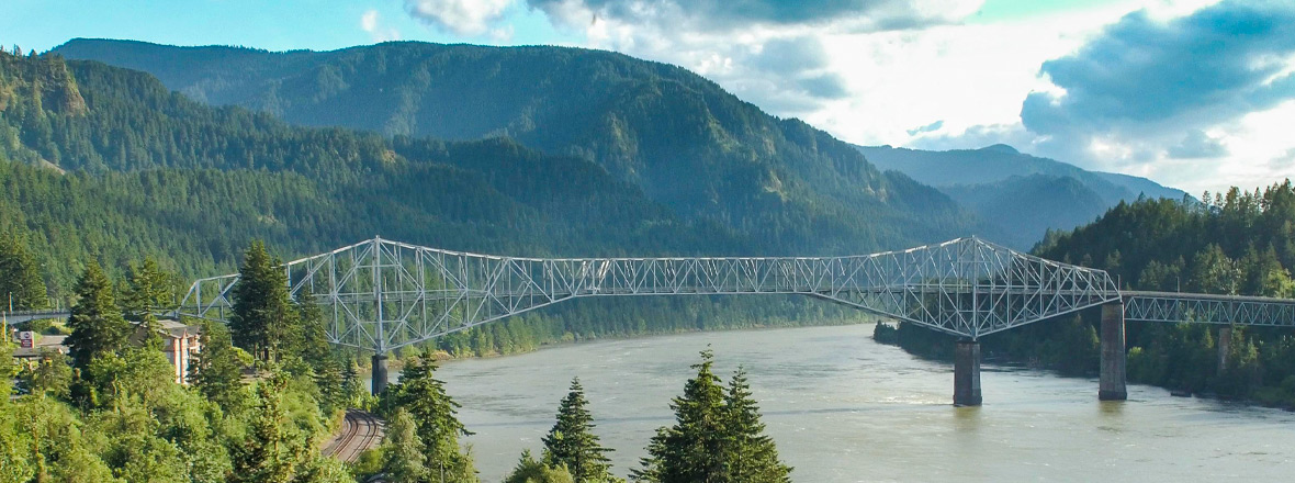 Bridge of the Gods, a steel truss cantilever bridge, spanning across the Columbia River. Evergreens surround the landscape.