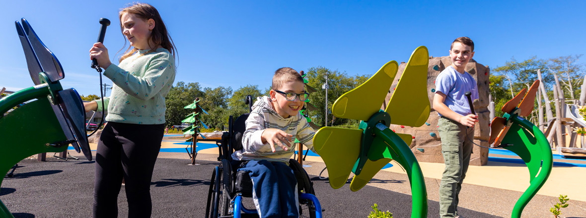 Three children, one in a wheelchair, play with accessible play structures.