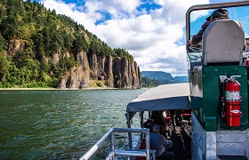 Boat traveling along a river past towering basalt cliffs and forested hills.