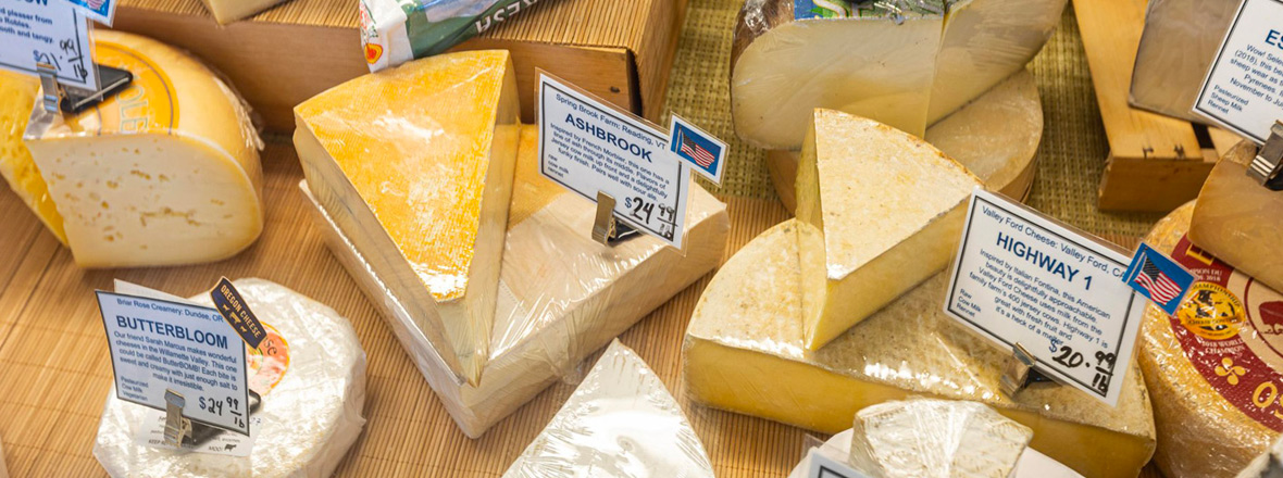 Display case filled with wedges and wheels of artisan cheese labeled with product names.