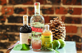 Bottles of spirits and a cocktail garnished with citrus, displayed alongside limes and a pinecone on a tabletop.
