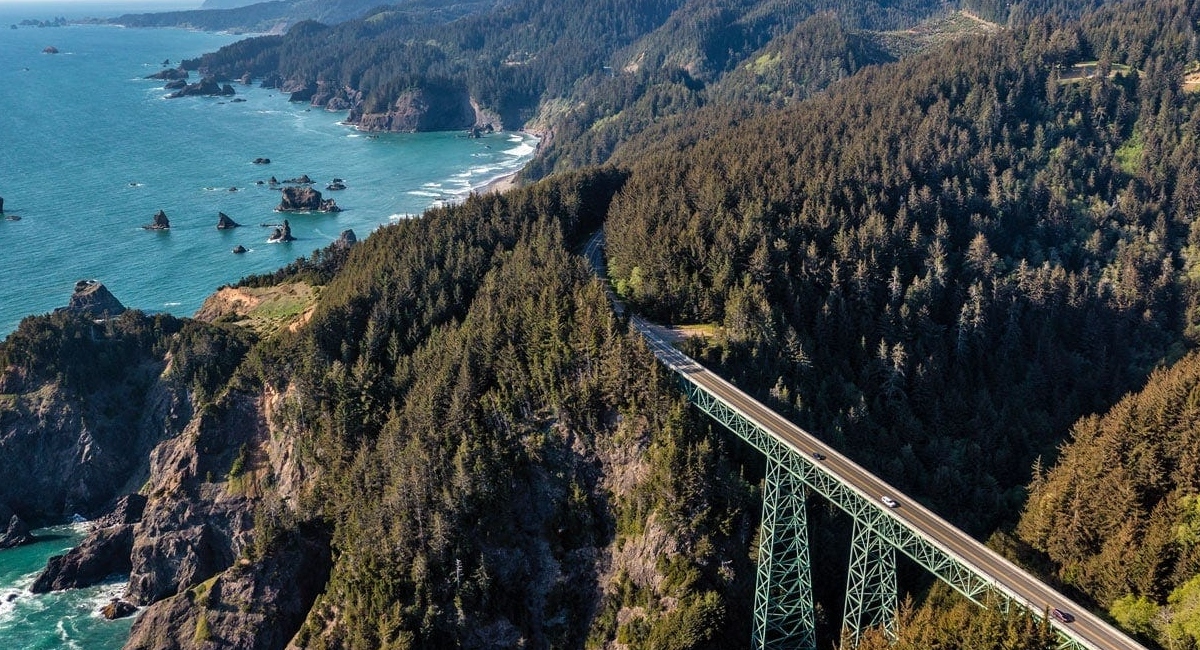 Aerial view of a coastal highway bridge spanning a forested canyon above the rugged Oregon coastline.