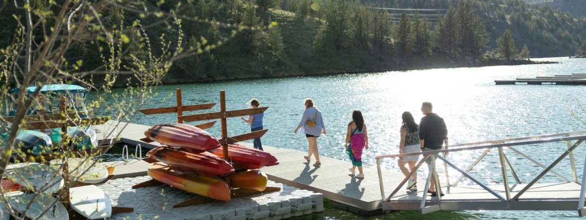 Five people walk down a walkway to a floating marina where a small boat is parked and several red kayaks and paddleboards are stacked. Trees and mountains are in the background and the lake is blue and sunny.