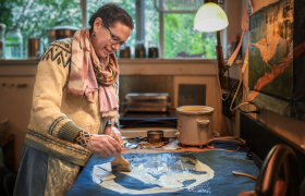 Person painting at an art studio table with brushes, paints and a landscape artwork in progress.