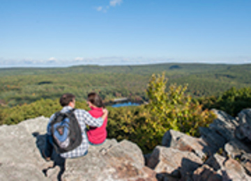Man with his arm around a woman sitting on a rock overlook admiring a lake surrounded by trees