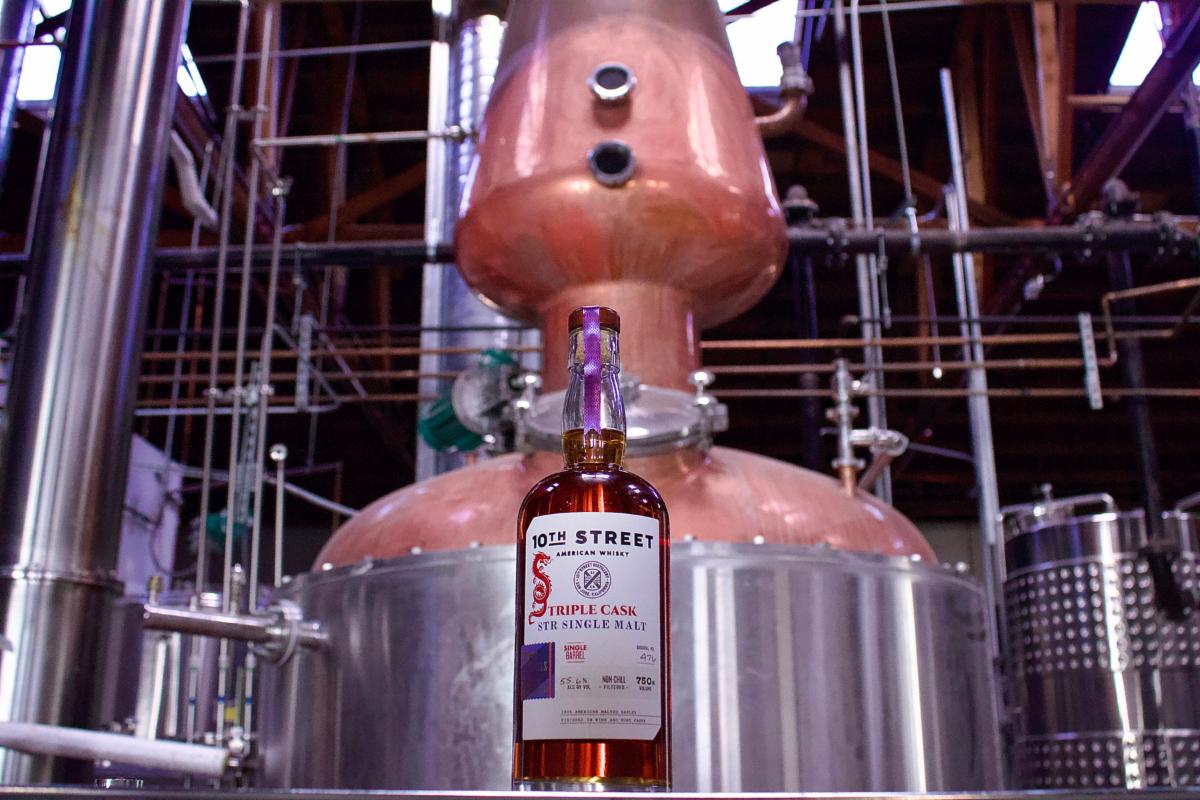 A bottle of 10th Street Distillery whiskey in front of a large distilling vat