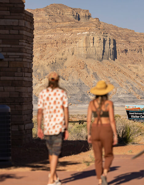a man and a woman walking down a dirt road with a rock formation in the background