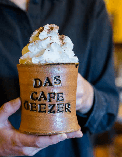 a mug that says "das cafe geezer" topped with a swirl of whipped cream