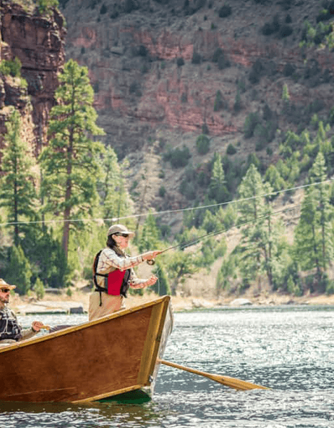 a woman flyfishing in a boat on a lake with a mountain in the background