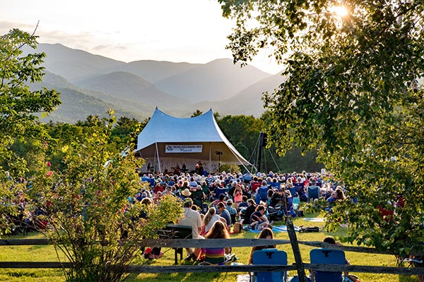 A crowd at a concert under a white tent with the sun peeking over mountain tops in the background.&nbsp;