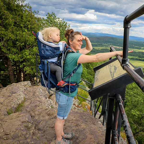 A person with a baby on their back takes in the view at a scenic overlook with mountains and a lake visible in the distance. 