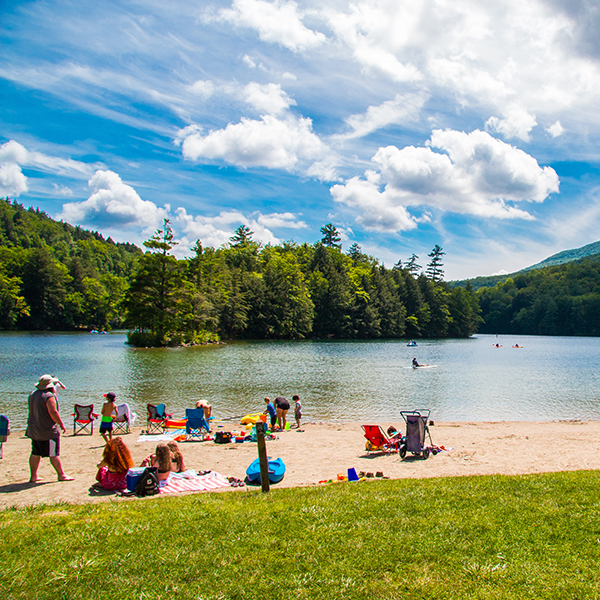  People swim and paddle in a blue lake or play or relax on a sandy beach on a sunny day. Trees are visible on the other side of the lake.