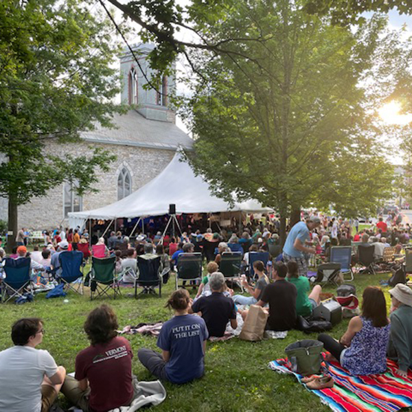 People sit outside on a summer day taking in live music with a grey stone church in the background.&nbsp;