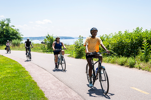 People riding bicycles on a path lined with green grass and bushes, with water visible in the background.