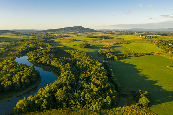 Aerial view of a lush green landscape with a winding river, dense forests, and expansive fields, under a clear sky with a distant mountain range.