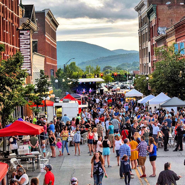 Crowds gather among white event tents at an outdoor festival in a historic downtown featuring brick buildings. 