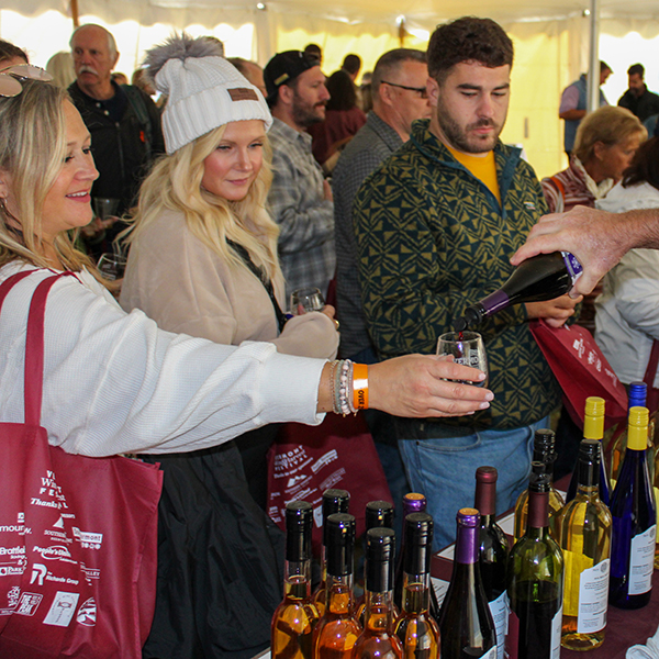 A person behind a table pours wine for people at the table at an outdoor festival under a tent.&nbsp;