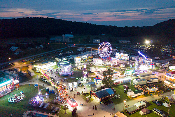 Aerial view of a lit-up carnival at dusk, featuring various rides, booths, and a Ferris wheel, with surrounding hills and a parking area in the background.