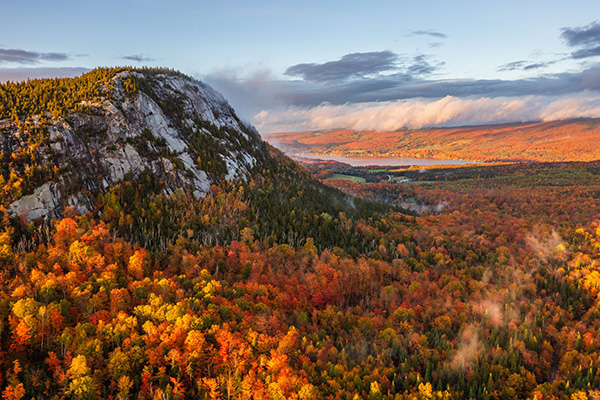 A tree-covered mountain with fall foliage on the trees, rendering the hillside bright orange, yellow, and red.
