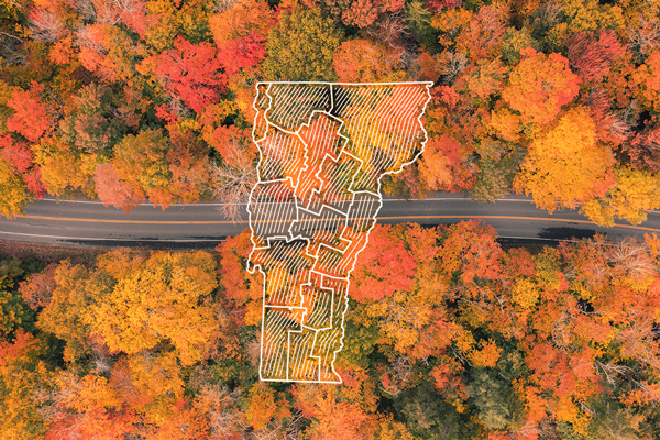 A road seen from far above passes by a lake and climbs a mountain in the fall with bright red and orange leaves on the trees.&nbsp;