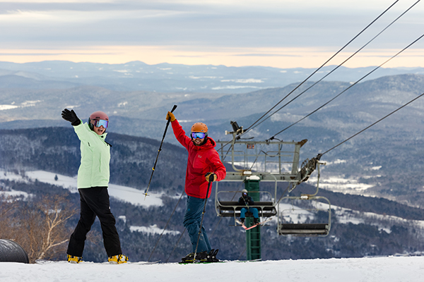 Two people at the top of a ski trail wave at the camera on a sunny day while a third person rides a chairlift behind them.&nbsp;