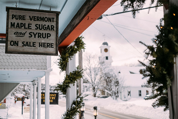 A sign reading “Pure Vermont Maple Sugar and Syrup On Sale Here” outside at a country store. Snow is visible behind. 