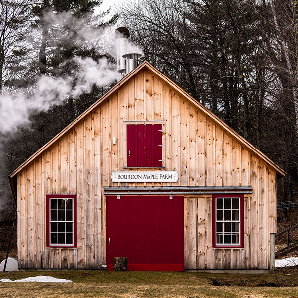 Seen from outside, a wooden sugarhouse in the spring with some snow still visible and steam coming from the top.