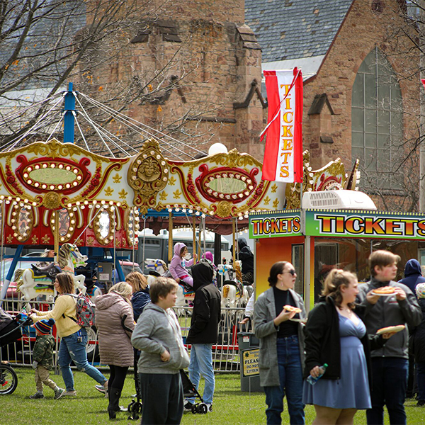 People seen outdoors at a street festival with historic buildings and carnival rides set up.&nbsp;