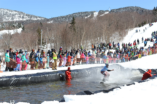 People watch as a person wearing a colorful costume on a snowboard splashes into a man-made pond at a snowy mountainside. 