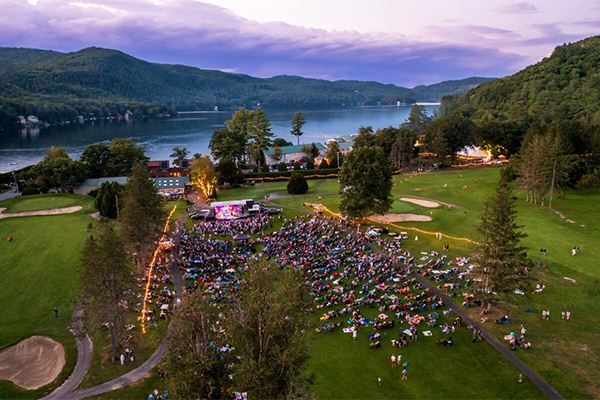Seen from far above, a crowd of people gather outside for a live musical performance with a lake and mountains in the background in the summer.&nbsp;