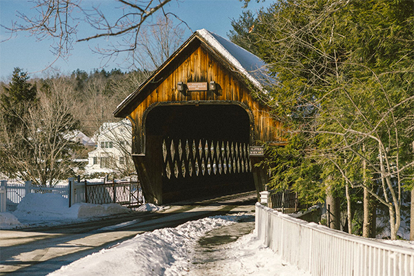  A wooden covered bridge and a white fence with snow around on a sunny winter day. 