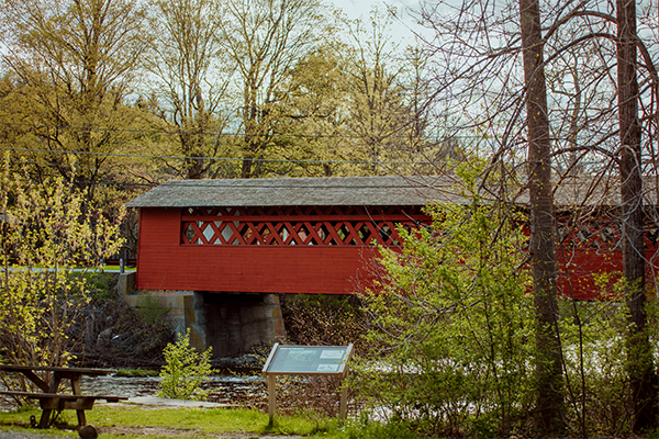 A red covered bridge with lattice framing surrounded by budding trees in the spring.&nbsp;