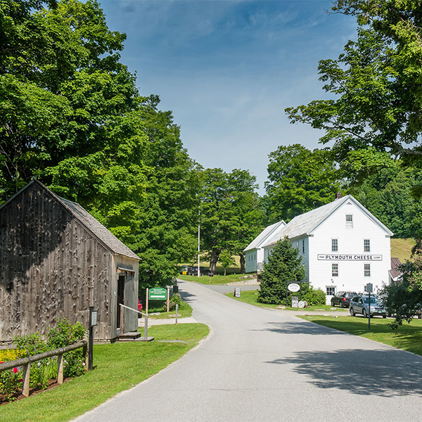A road curves past a weathered wooden shed and a white building labeled "Plymouth Cheese," surrounded by green trees and grass under a blue sky.