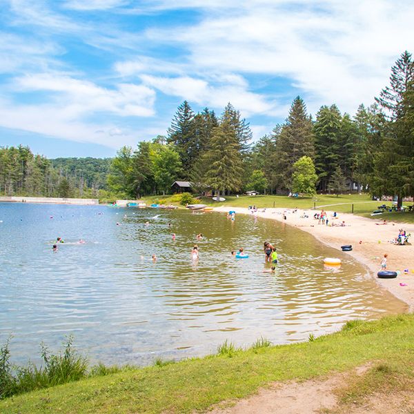 People splash and swim in a lake next to a sandy beach where others relax on a sunny summer day.