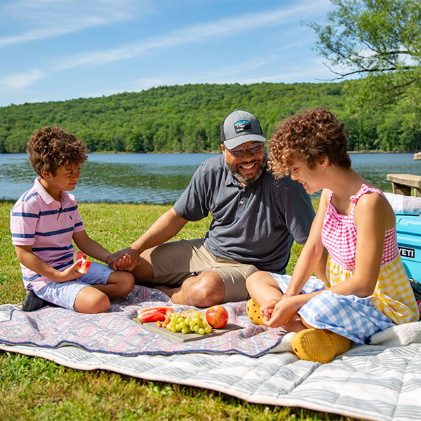 Three people are enjoying a picnic on a blanket in a grassy area near a body of water. 