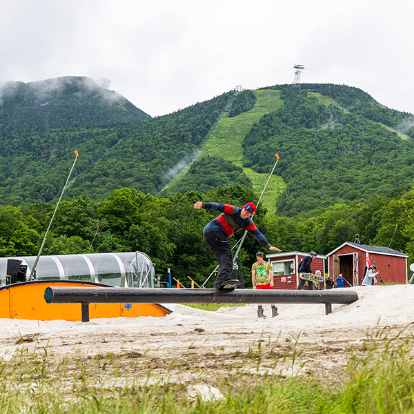  A person on a snowboard does tricks at a terrain park on a thin layer of snow with grass elsewhere and on the mountain behind.