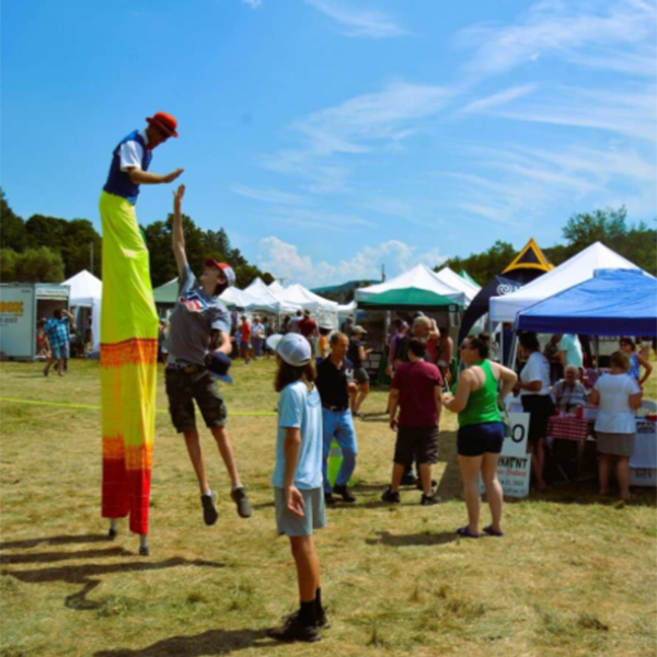 A stilt walker high-fives a person at an outdoor festival. Tents and booths are in the background, with people enjoying the sunny day.