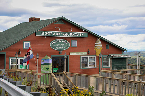 A red general store building seen from outside in the summer with grass growing and flowers blooming. Behind the building, mountains are visible.