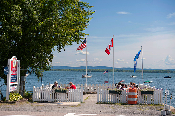 Various flags fly overhead on a summer day as people dine by a lake.