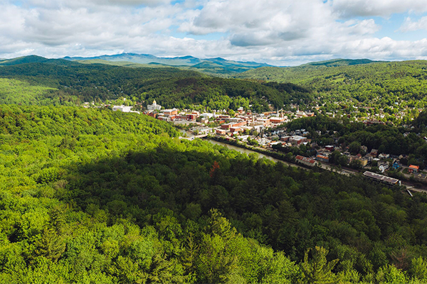 Aerial view of downtown Montpelier surrounded by lush green forests and distant mountains under a partly cloudy sky.