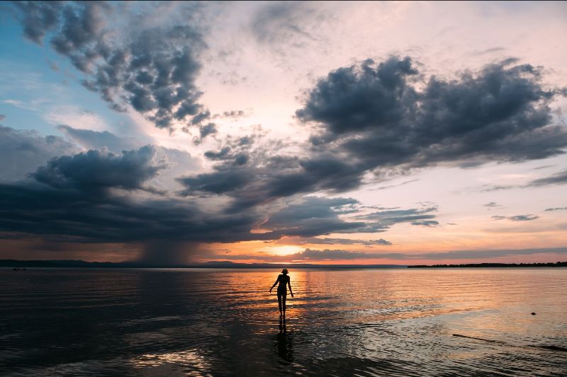 A person walks into a lake at sunset, seen from behind in silhouette.