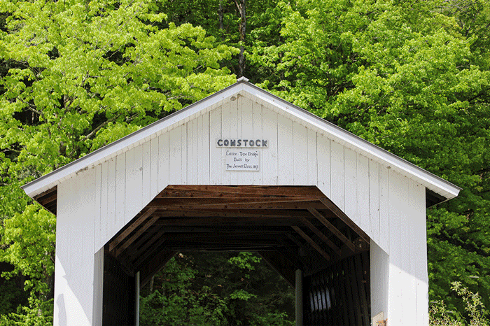 An animated GIF showcasing a series of clips of the six historic covered bridges in Montgomery, Vermont, known as the "Covered Bridge Capital of Vermont." The GIF transitions between the different bridges, including the Comstock Bridge, built in 1883, and other bridges like the Fuller, Hutchin's, and Longley bridges. The clips feature various angles of these wooden structures and highlight their rustic beauty crossing rivers, surrounded by lush trees with green leaves, and including some yellow and white road signs.