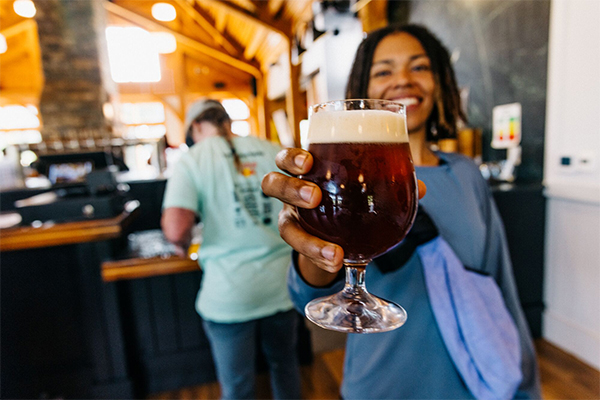 A person holds a full beer glass out to the camera, smiling.