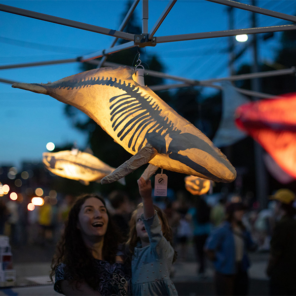 At night an adult and a child stop to admire a piece of lit art hanging above. 
