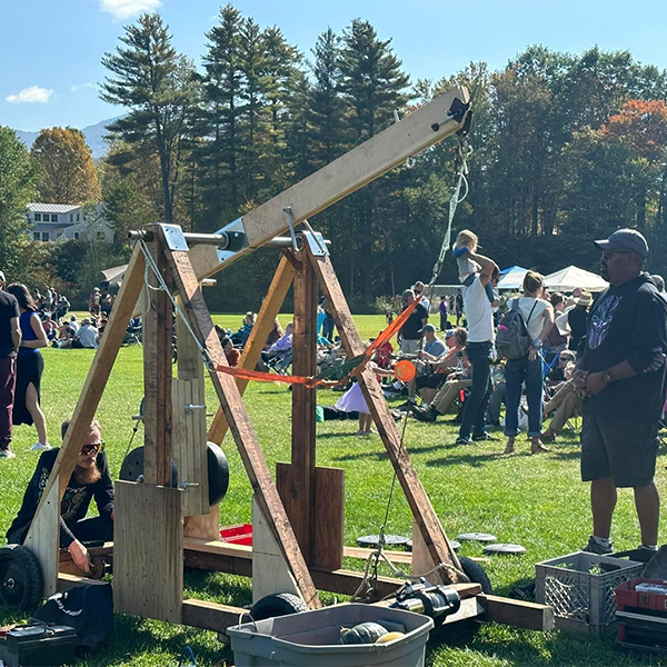 People gather around trebuchets made out of wood with pumpkins around ready to be launched on a sunny fall day.