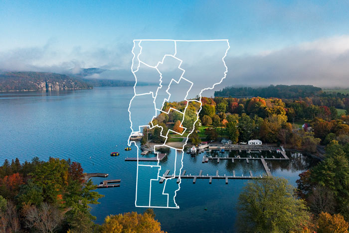 Aerial view of a Vermont lake with marina docks and colorful autumn foliage, featuring a white outline of the Vermont state map with county boundaries.